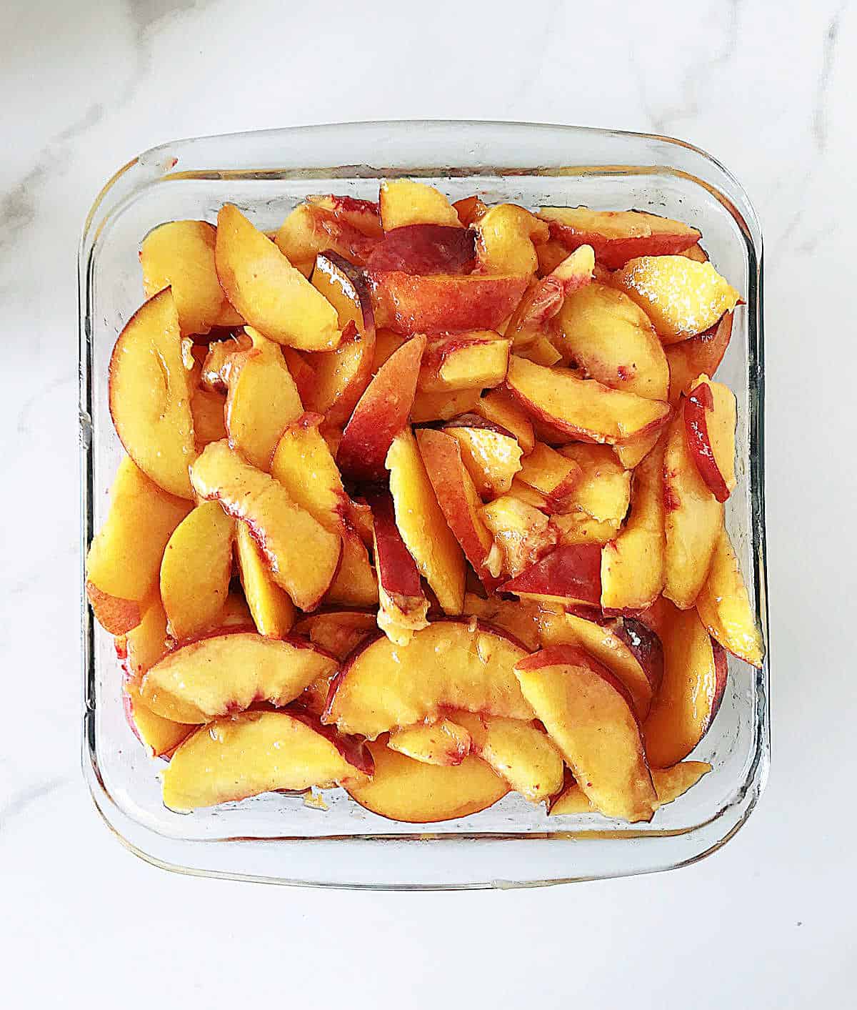 Square glass dish with sliced peaches on marble counter.