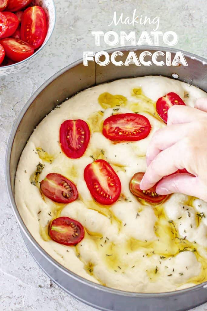 Hand adding tomatoes to focaccia dough in round pan, grey surface, white text overlay