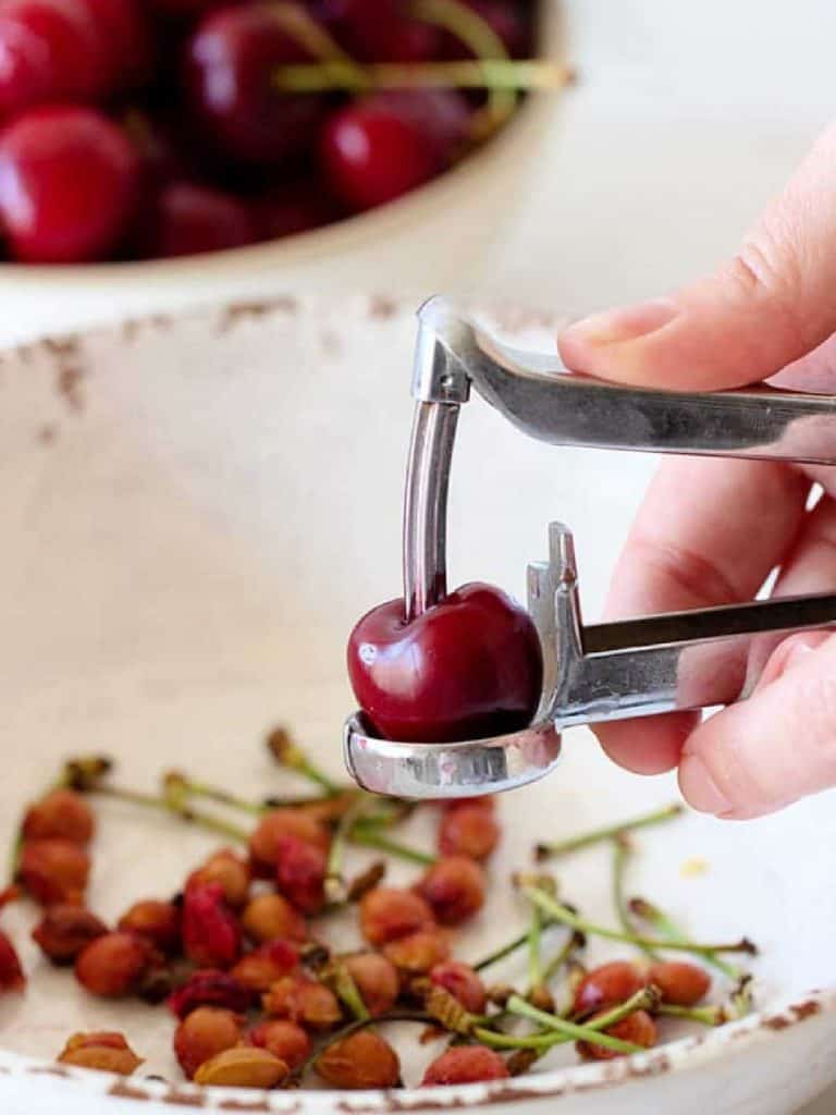 A fresh cherry being pitted over a white bowl with pits.