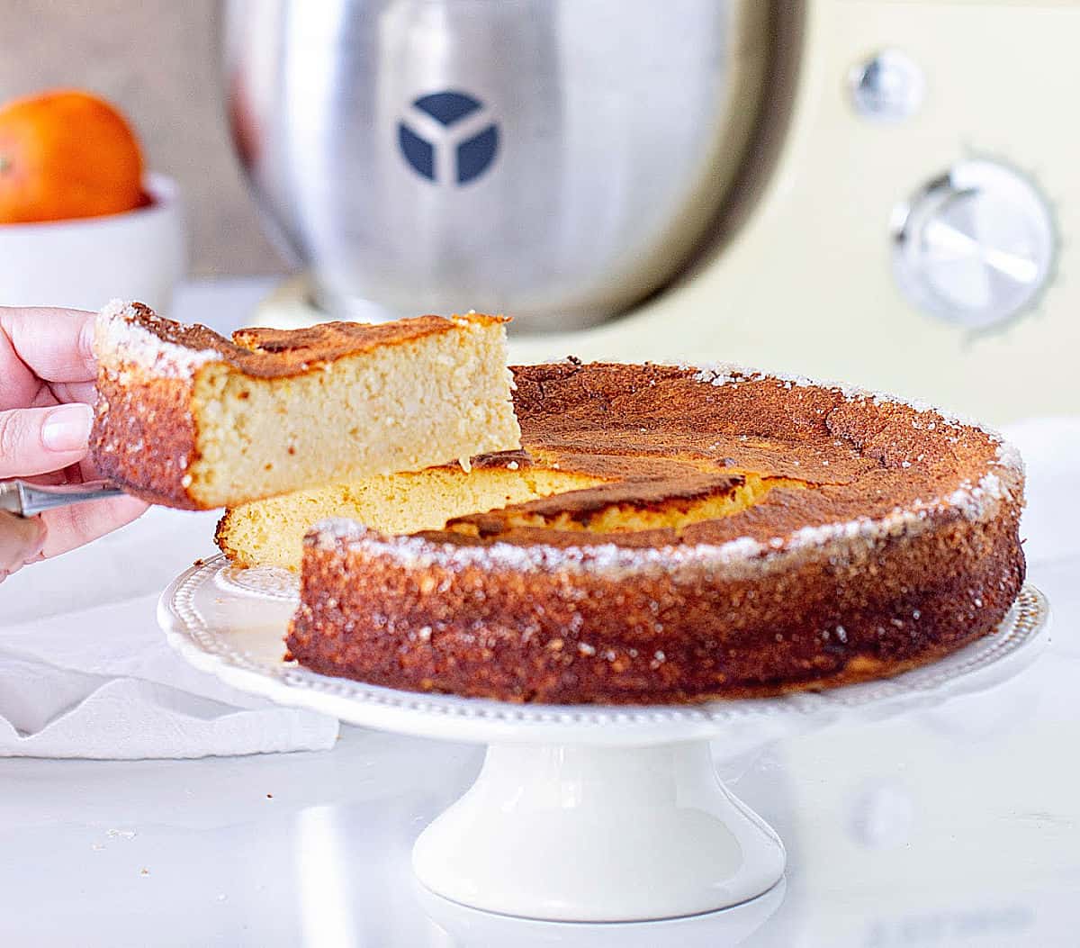 Ricotta Cheesecake on a cake stand, hand holding a slice, white surface, stand mixer in the backgorund