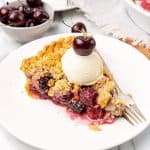 Ice cream topped cherry crumb pie in a white plate. A fork, bowl of cherries.