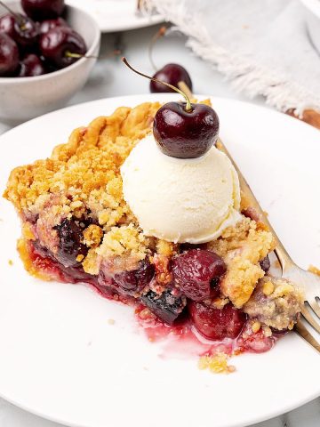 Ice cream topped cherry crumb pie in a white plate. A fork, bowl of cherries.