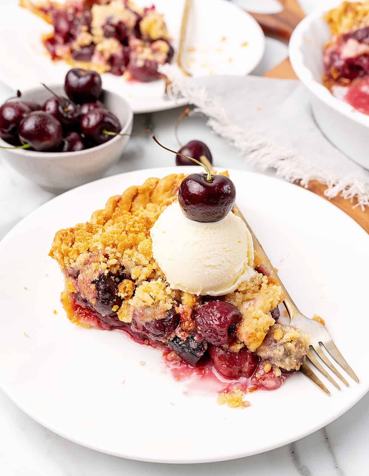 White plate with cherry crumb pie slice with ice cream, bowl with cherries and more pie in the background.