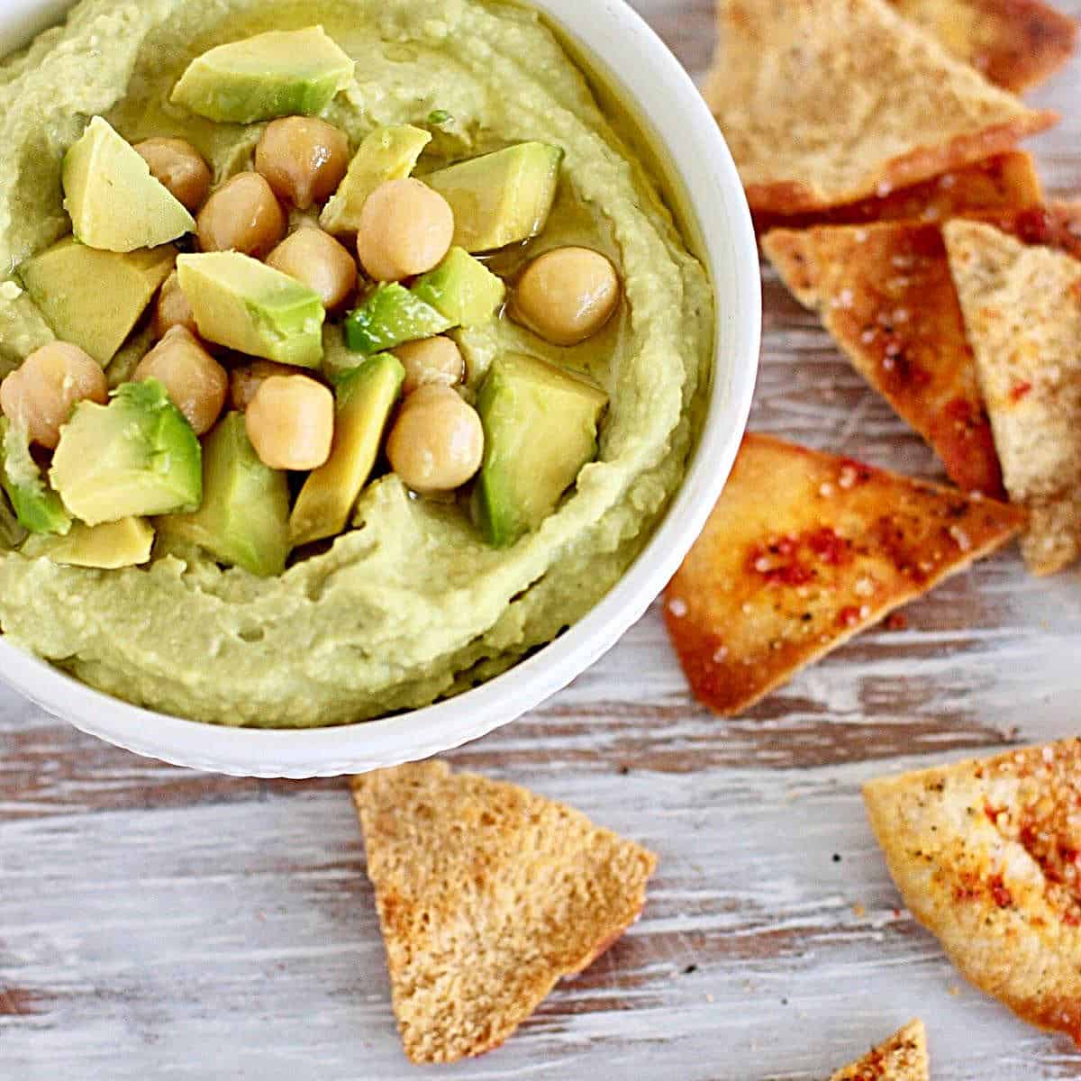 Avocado hummus dip in a white bowl with chickpeas, avocado dice, and pita chips. White wooden surface.