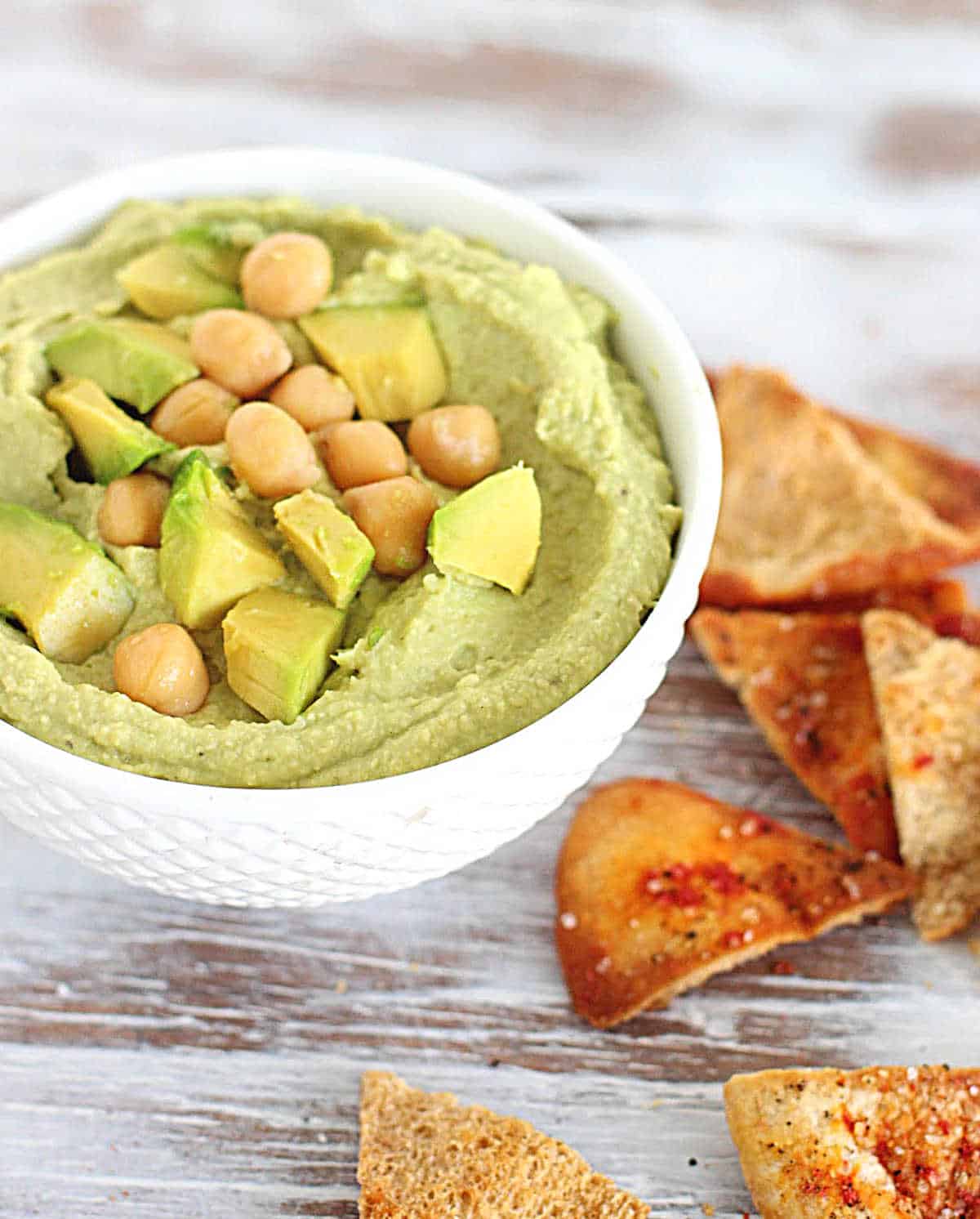 White bowl with green dip, chickpeas and pita chips; on a white wooden table.