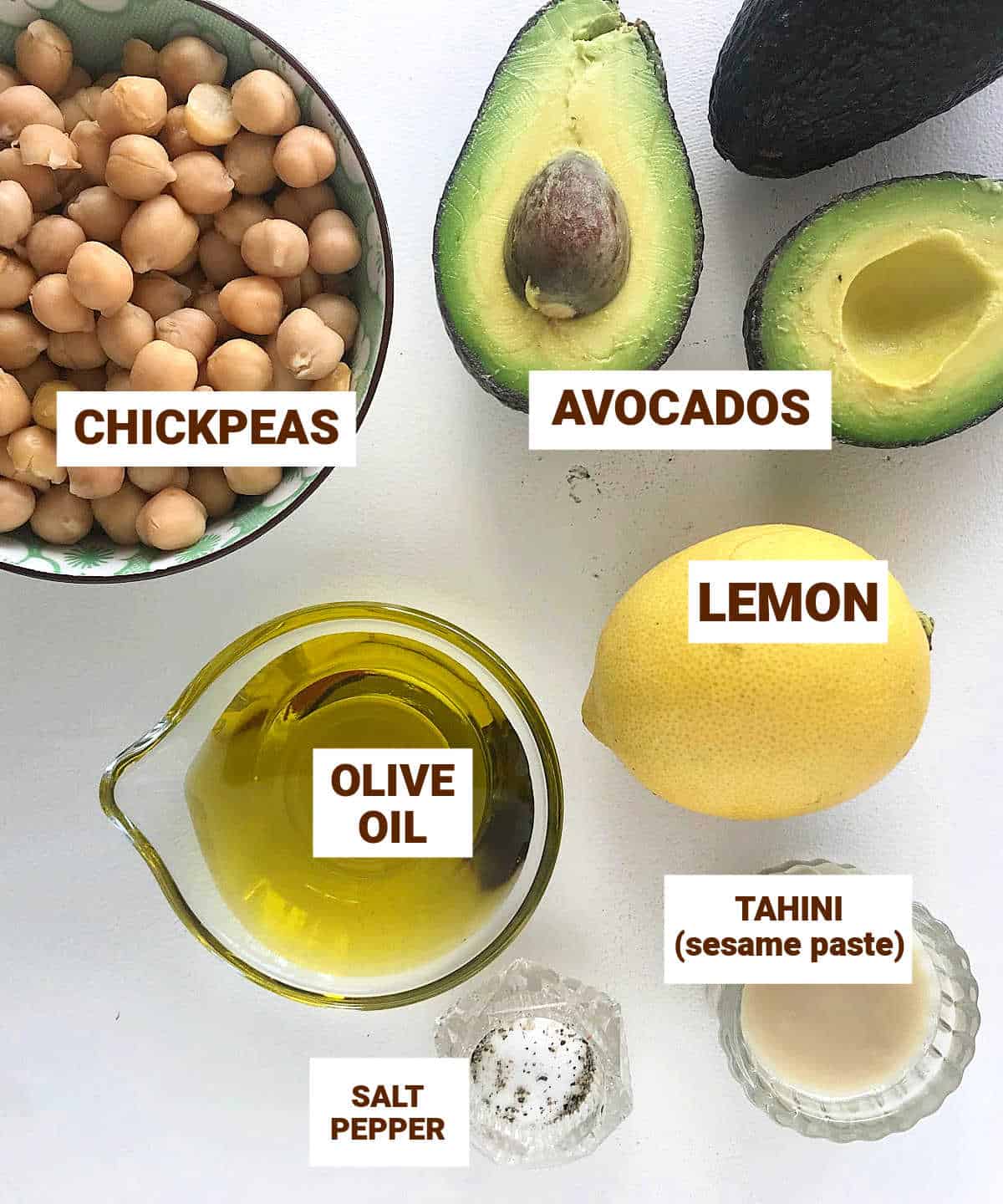 Ingredients for avocado hummus , in bowls on white surface, including lemon, chickpeas, oil, tahini.