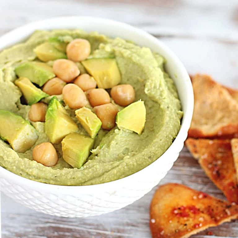 Close up of white bowl with avocado dip on a white surface with pita chips.
