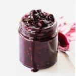 Glass jar with blueberry sauce, a spoon, white background.