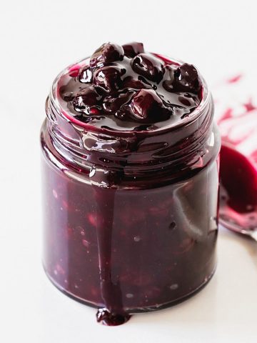 Glass jar with blueberry sauce, a spoon, white background.
