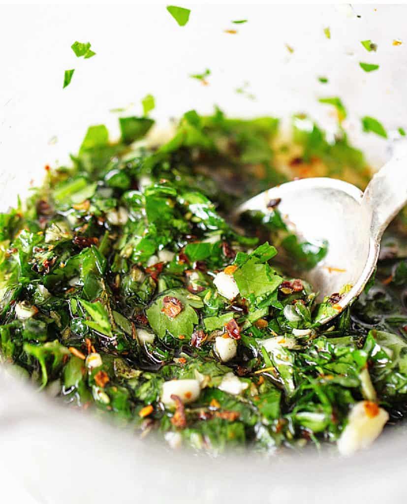 A glass jar with chimichurri sauce, a silver spoon, close up image