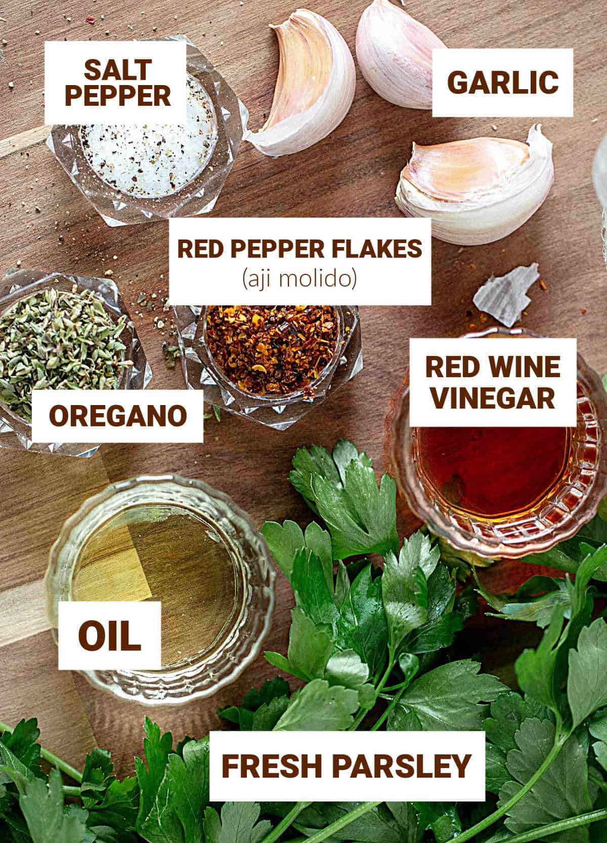 Garlic cloves, fresh parsley and rest of chimichurri ingredients in small bowls on wooden board, text overlay