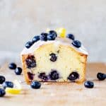 Square image of cut blueberry bread on wooden board, grey background, loose fresh blueberries