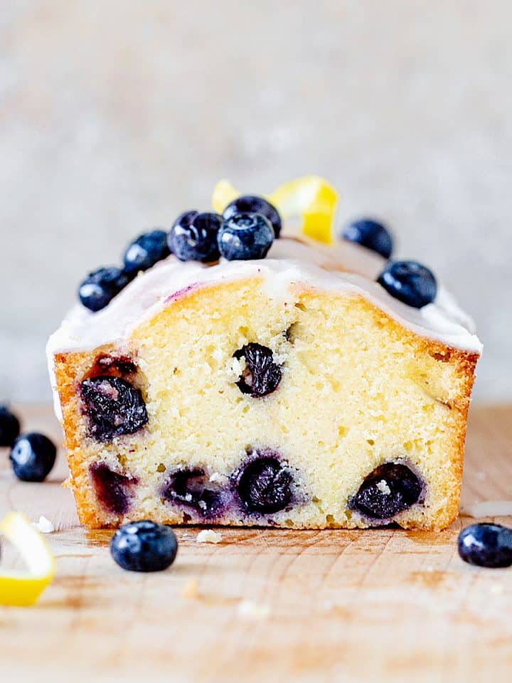 Square image of cut blueberry bread on wooden board, grey background, loose fresh blueberries