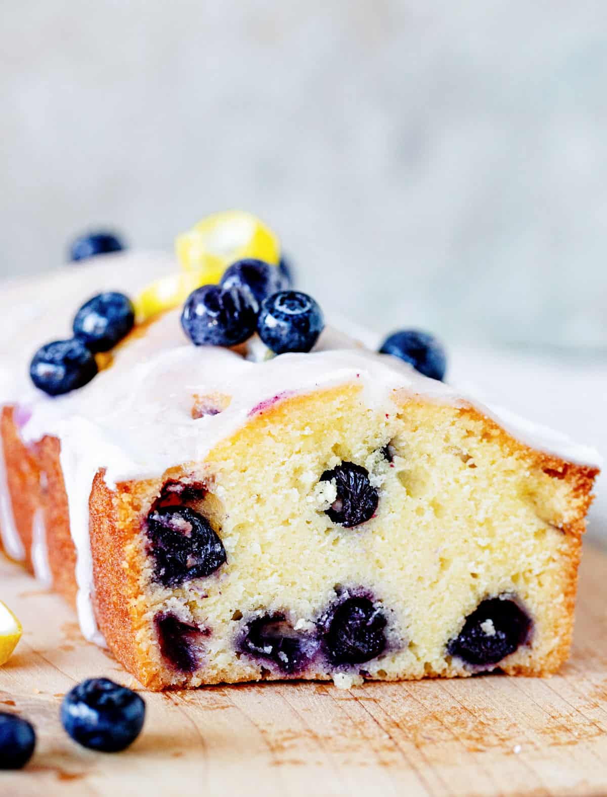 Glazed blueberry pound cake with front cut on wooden board, blueberries around, grey background.
