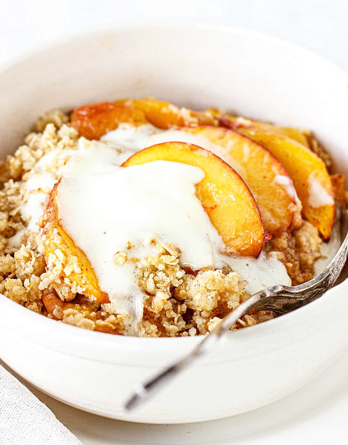 Peach crisp serving with ice cream in white bowl with a silver spoon.