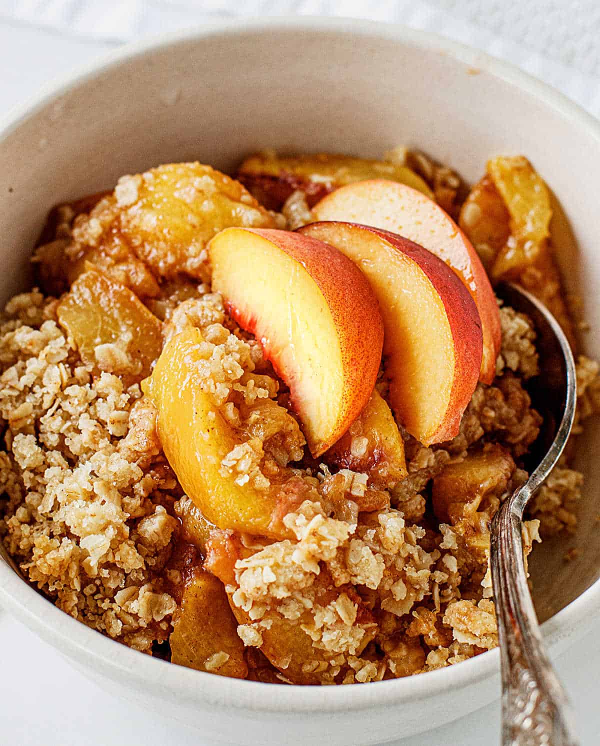 White bowl with peach crisp serving and silver spoon inside.