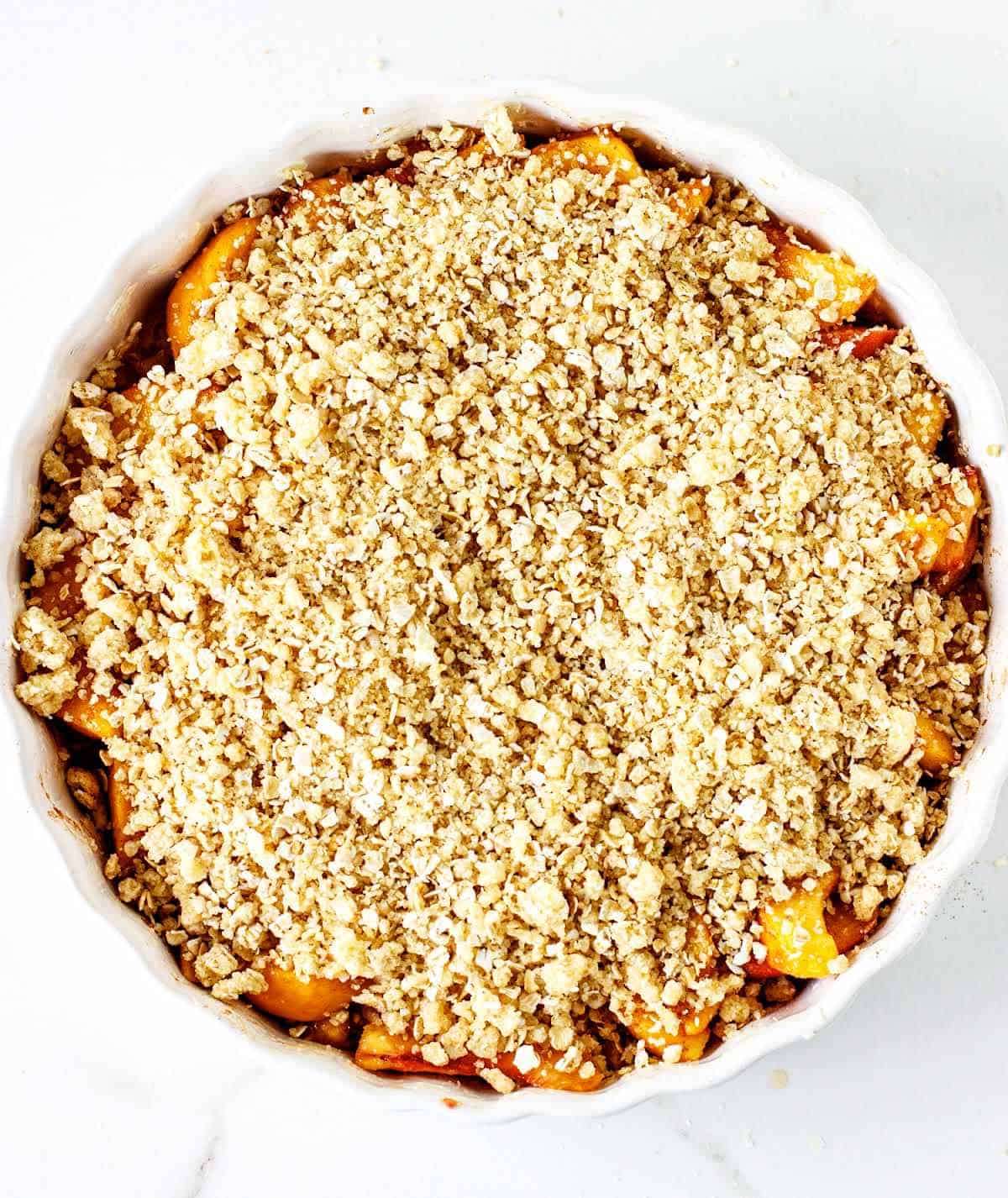 Round dish with unbaked peach crisp on white surface.