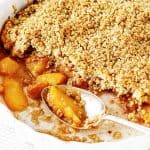 A white round dish with baked peach crumble and a silver spoon.