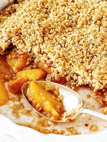 A white round dish with baked peach crumble and a silver spoon.