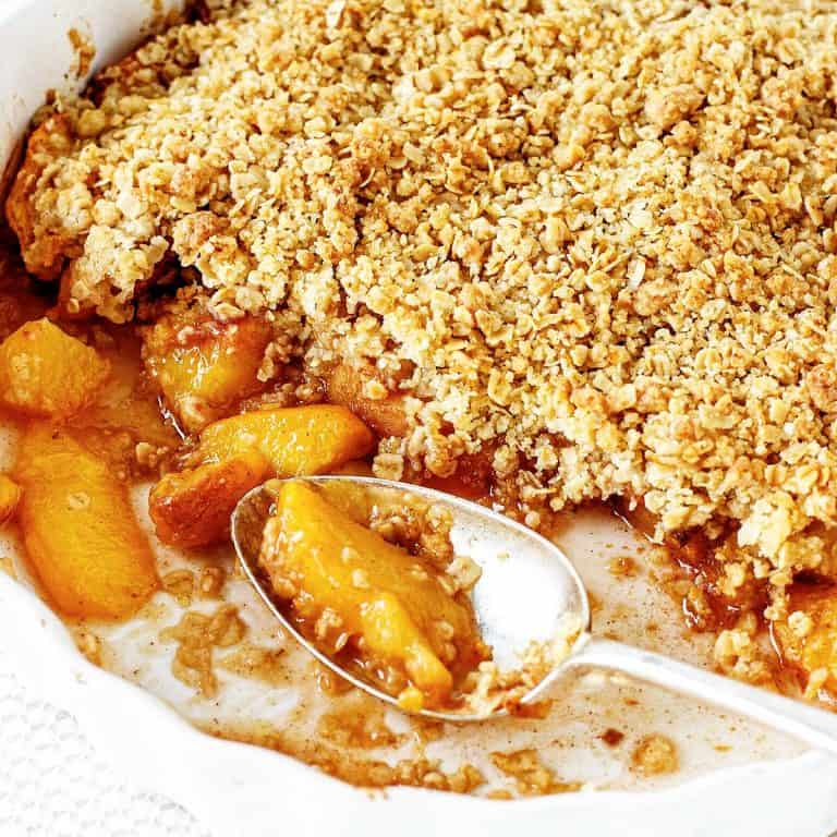 A white round dish with baked peach crumble and a silver spoon.