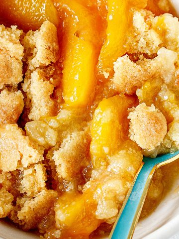 Close view of peach dump cake serving in white bowl with a teal spoon.
