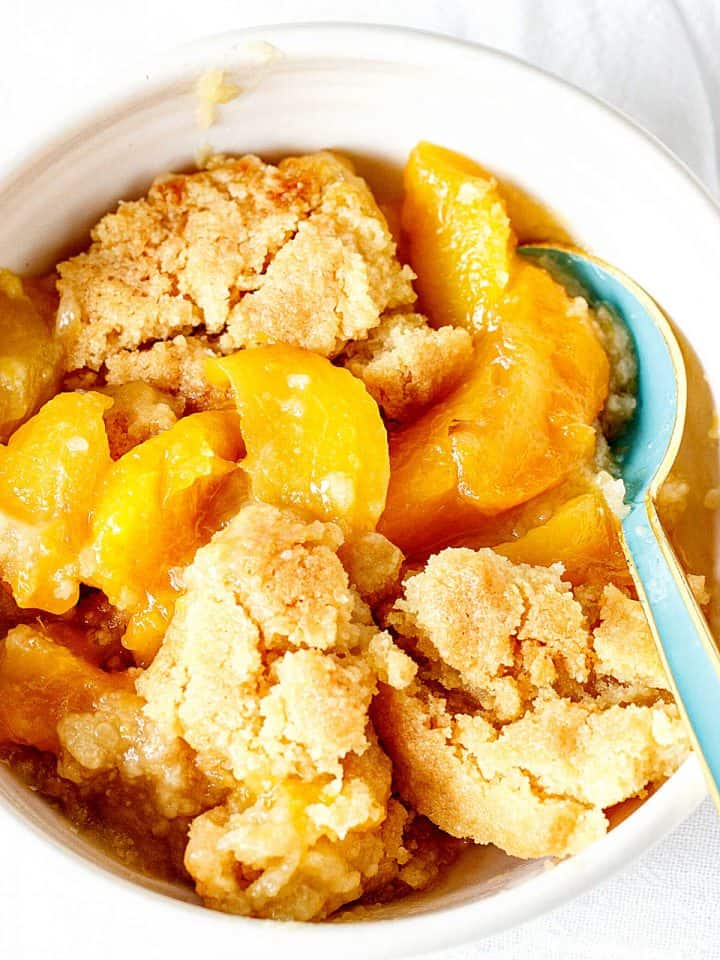 A white bowl with peach dump cake, blue spoon