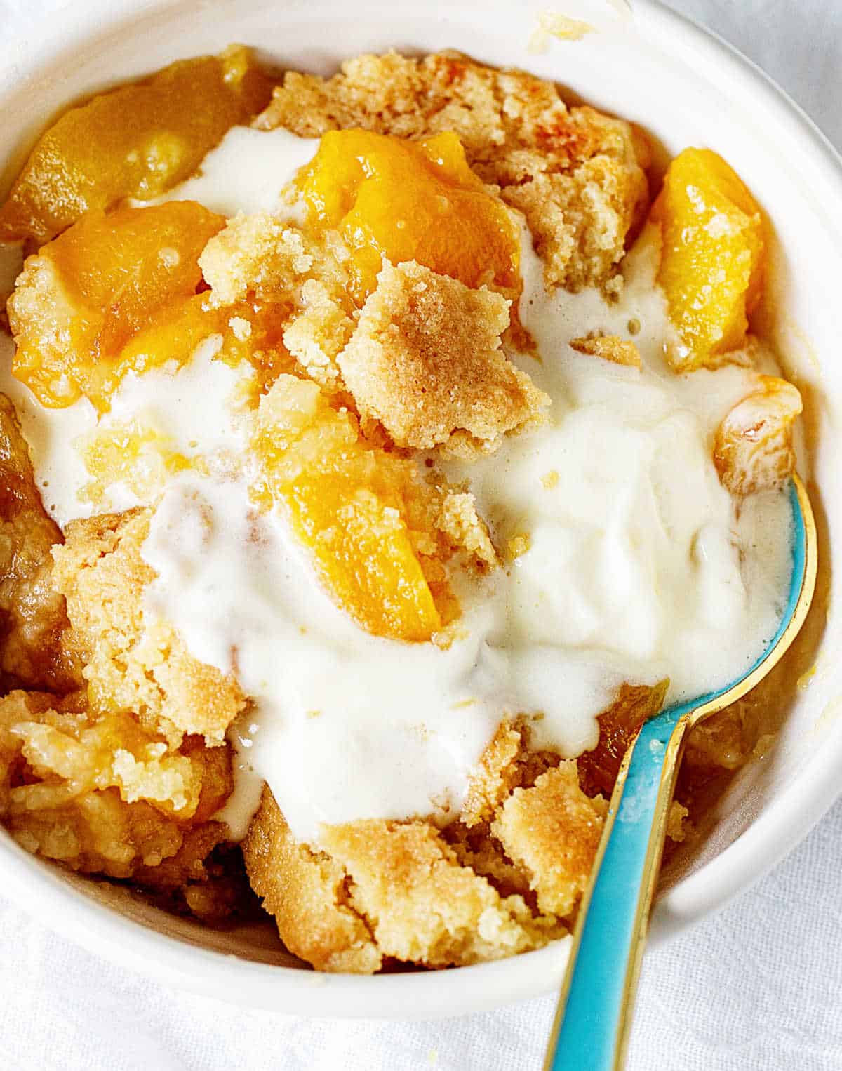 Overview of white bowl with peach dump cake and ice cream, greenish spoon