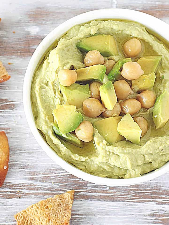 Washed white table with bowl containing avocado dip, chickpeas on top, pita chips around