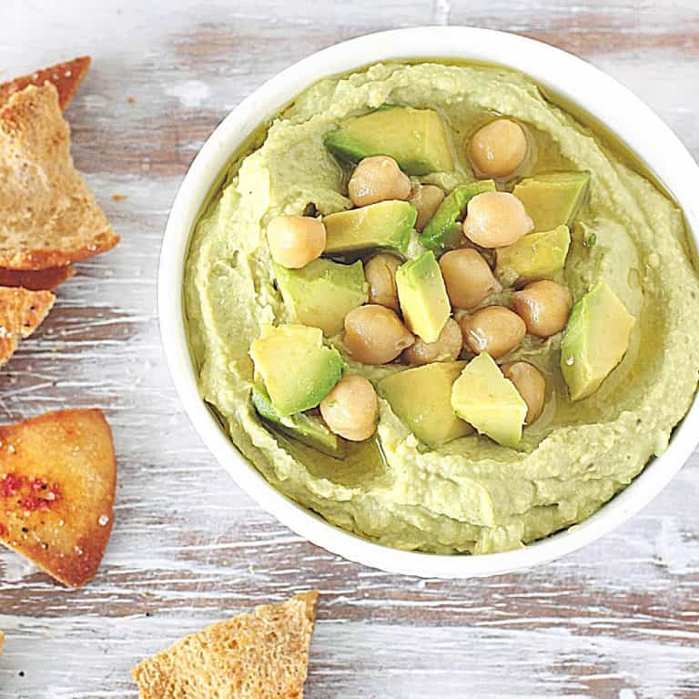 Washed white table with bowl containing avocado dip, chickpeas on top, pita chips around