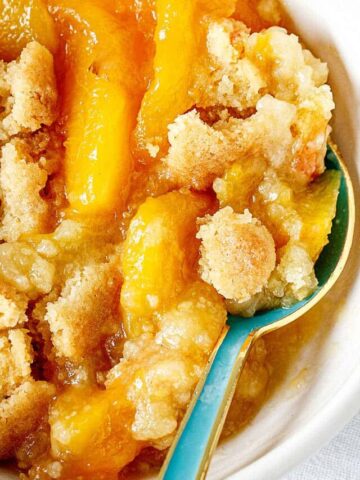 Close view of peach dump cake serving in white bowl with a teal spoon.