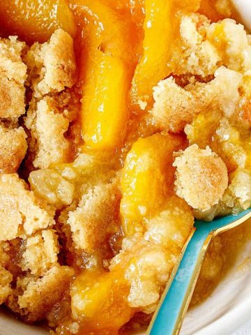 Close view of peach dump cake serving in white bowl with a teal spoon.