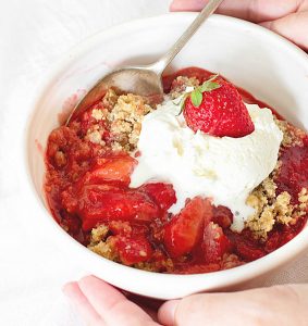 Strawberry crisp serving with spoon in white bowl, hands holding it