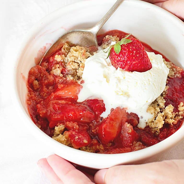 White bowl with strawberry crisp and ice cream, hand holding silver spoon