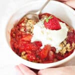 White bowl with serving of strawberry crisp with ice cream scoop. Silver spoon.