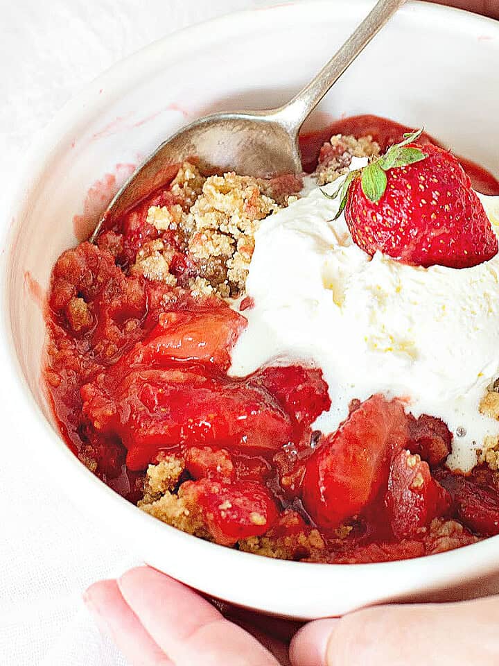 White bowl with serving of strawberry crisp with ice cream scoop. Silver spoon.