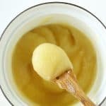 Spoon with applesauce over white saucepan, white surface, red text overlay