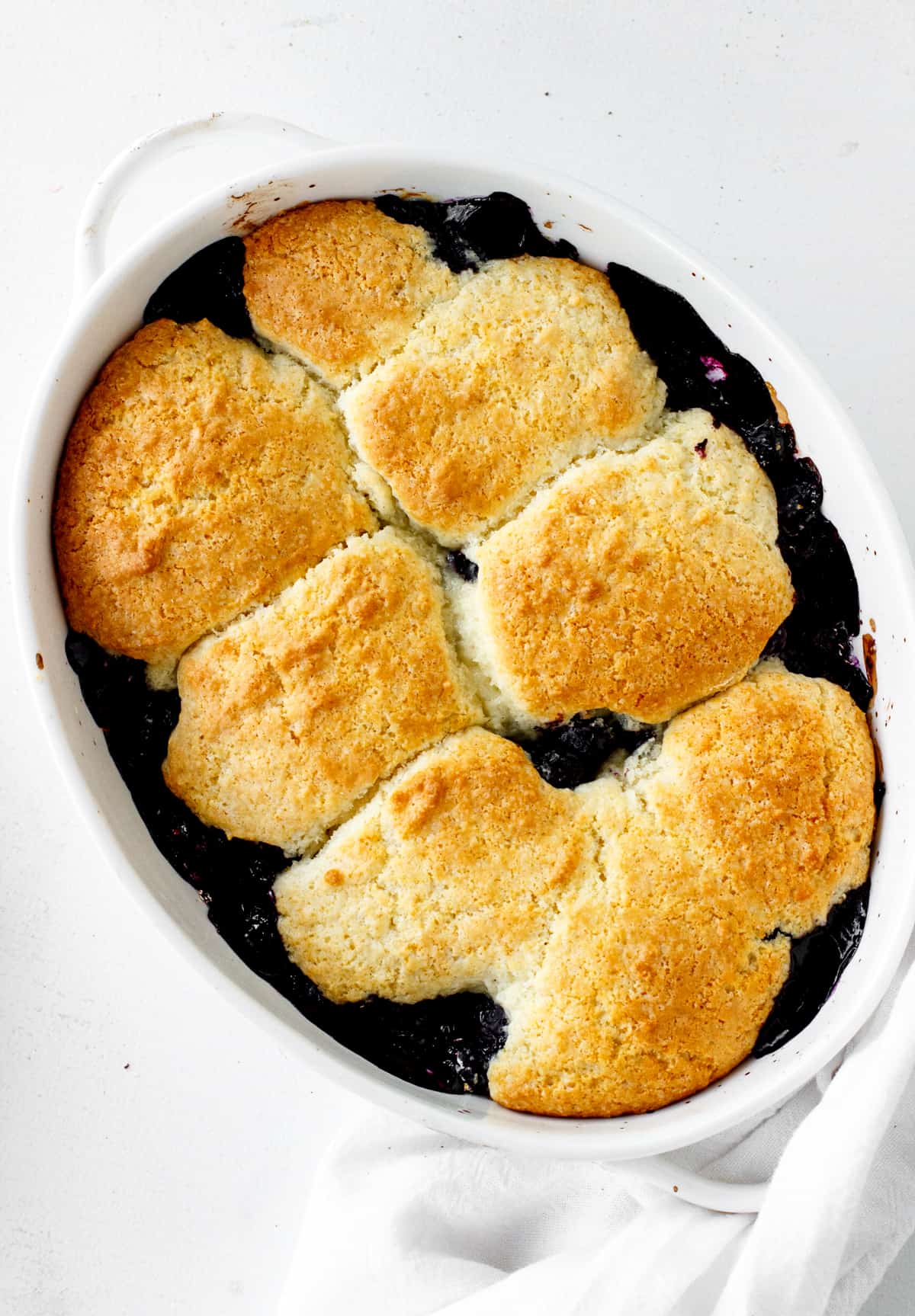 Overview of baked blueberry cobbler in oval dish on a white surface