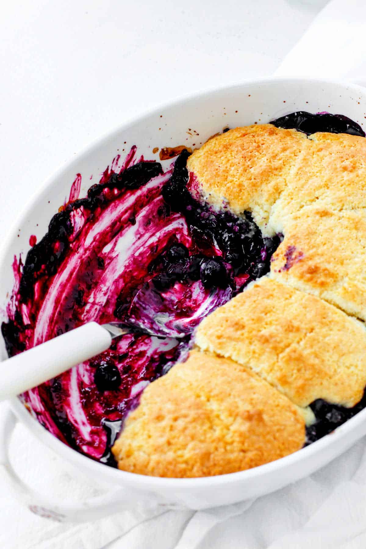 White dish with blueberry cobbler, spoon inside, white surface.