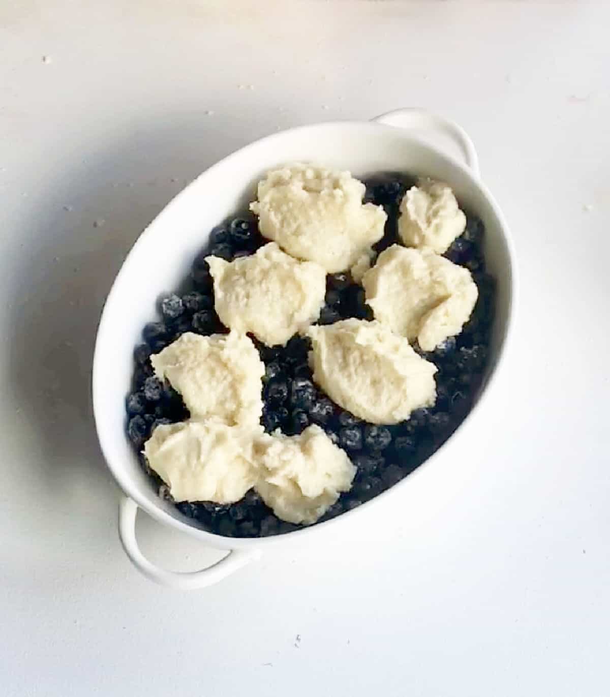 Top view of oval white dish with unbaked blueberry cobbler on a white surface.