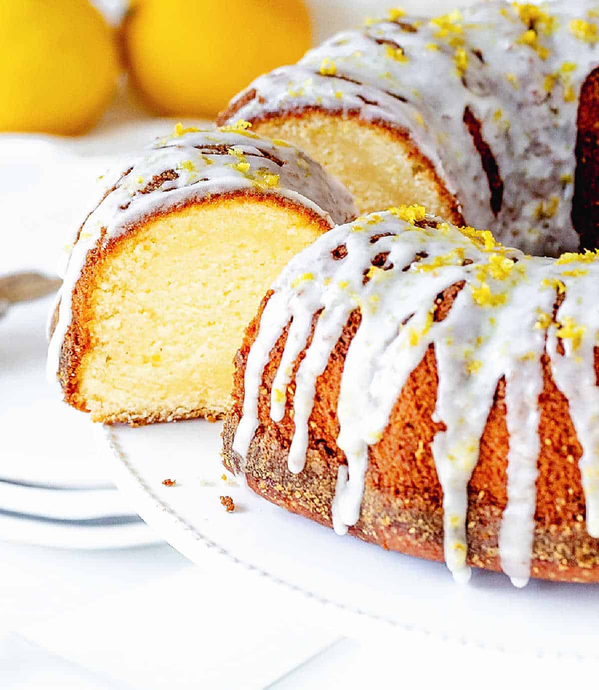 A slice of lemon bundt cake cut from cake in white stand, lemons in background