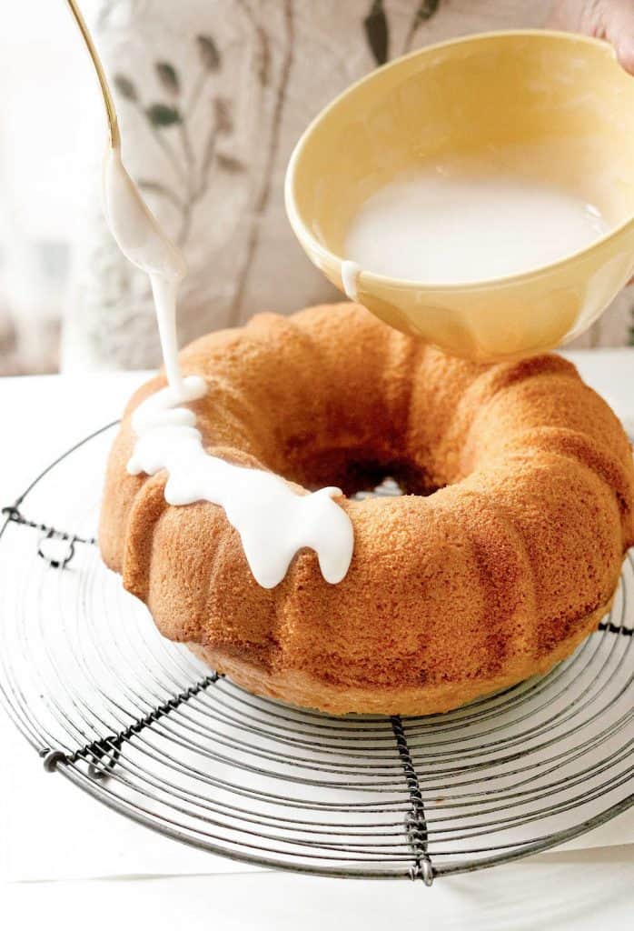 Drizzling lemon glaze from a yellow bowl onto a bundt cake on a wire rack.