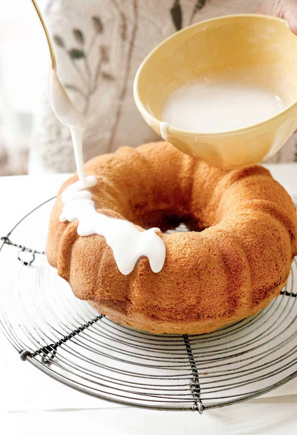 Glazing a bundt cake on a wire rack on a white surface. Yellow bowl with glaze.
