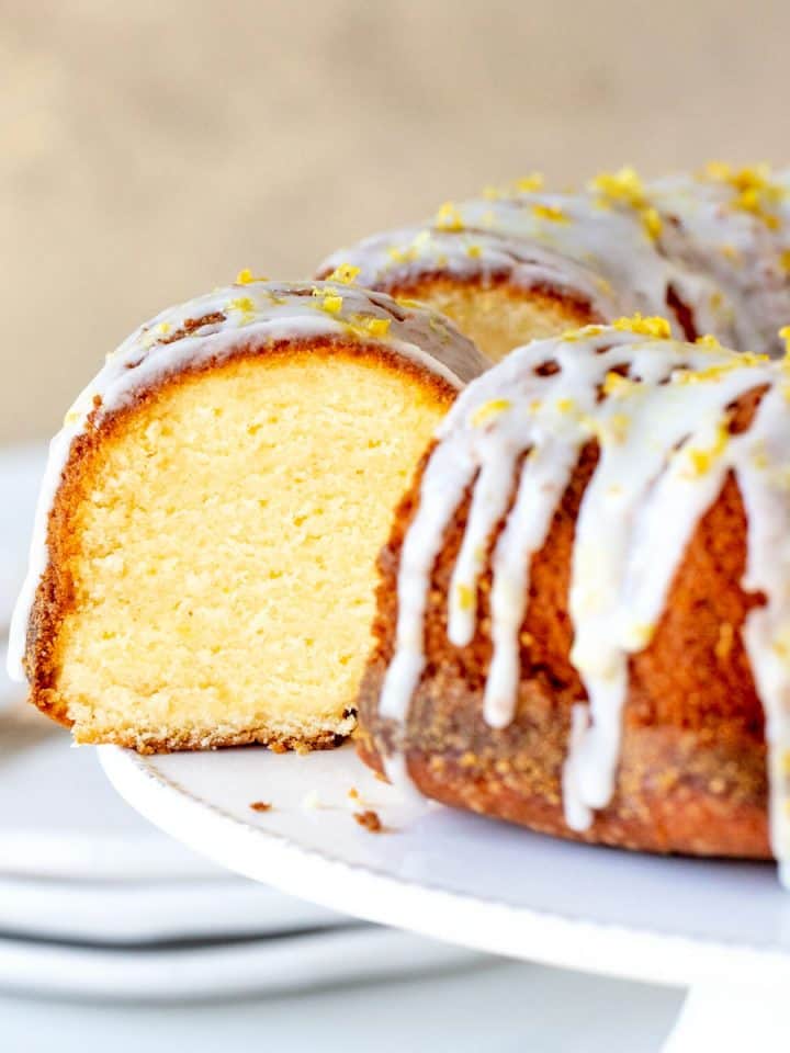Lemon bundt cake with glaze and cut slice on white cake stand, beige background.
