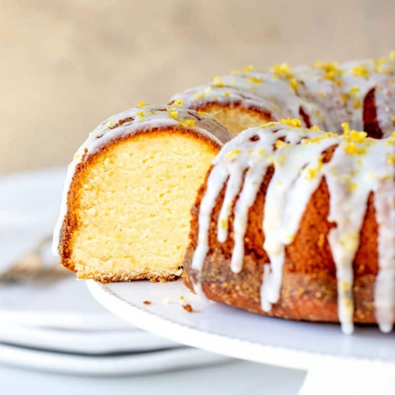 Lemon bundt cake with glaze and cut slice on white cake stand, beige background.