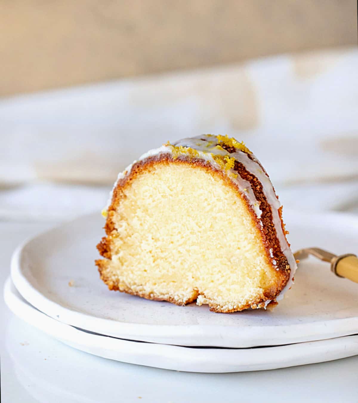 Single slice of glazed lemon bundt cake on stack of white plates. White and beige background. 