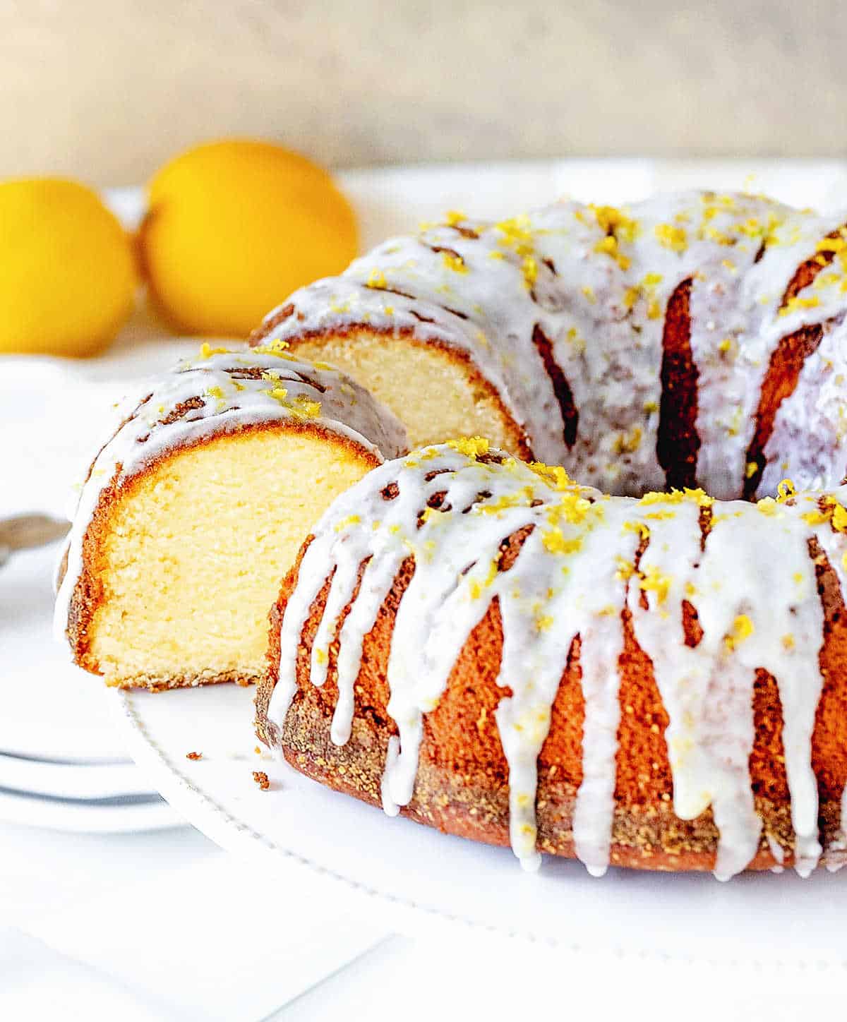 Glazed lemon bundt cake with cut slice on a white stand, whole lemons, white beige background.