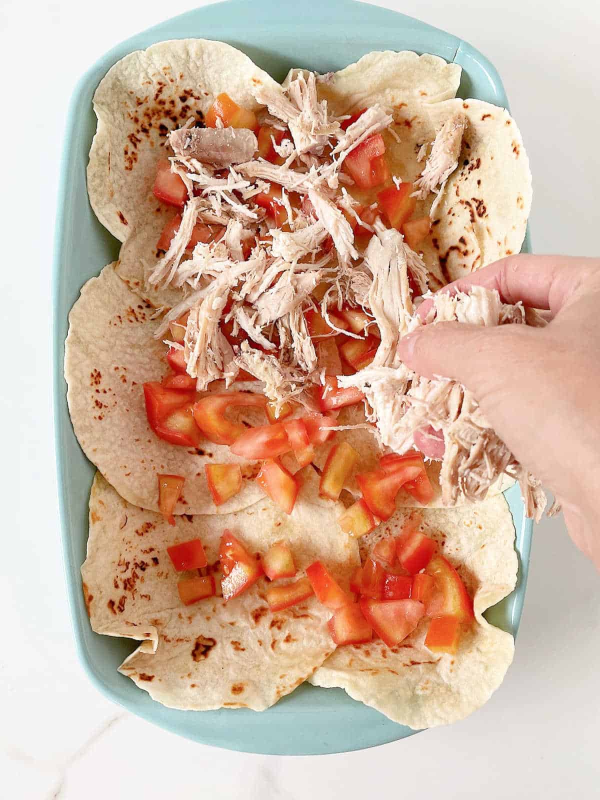 Hand adding chicken and tomatoes on a layer of tortillas in a rectangular blue baking dish on white marble surface. 