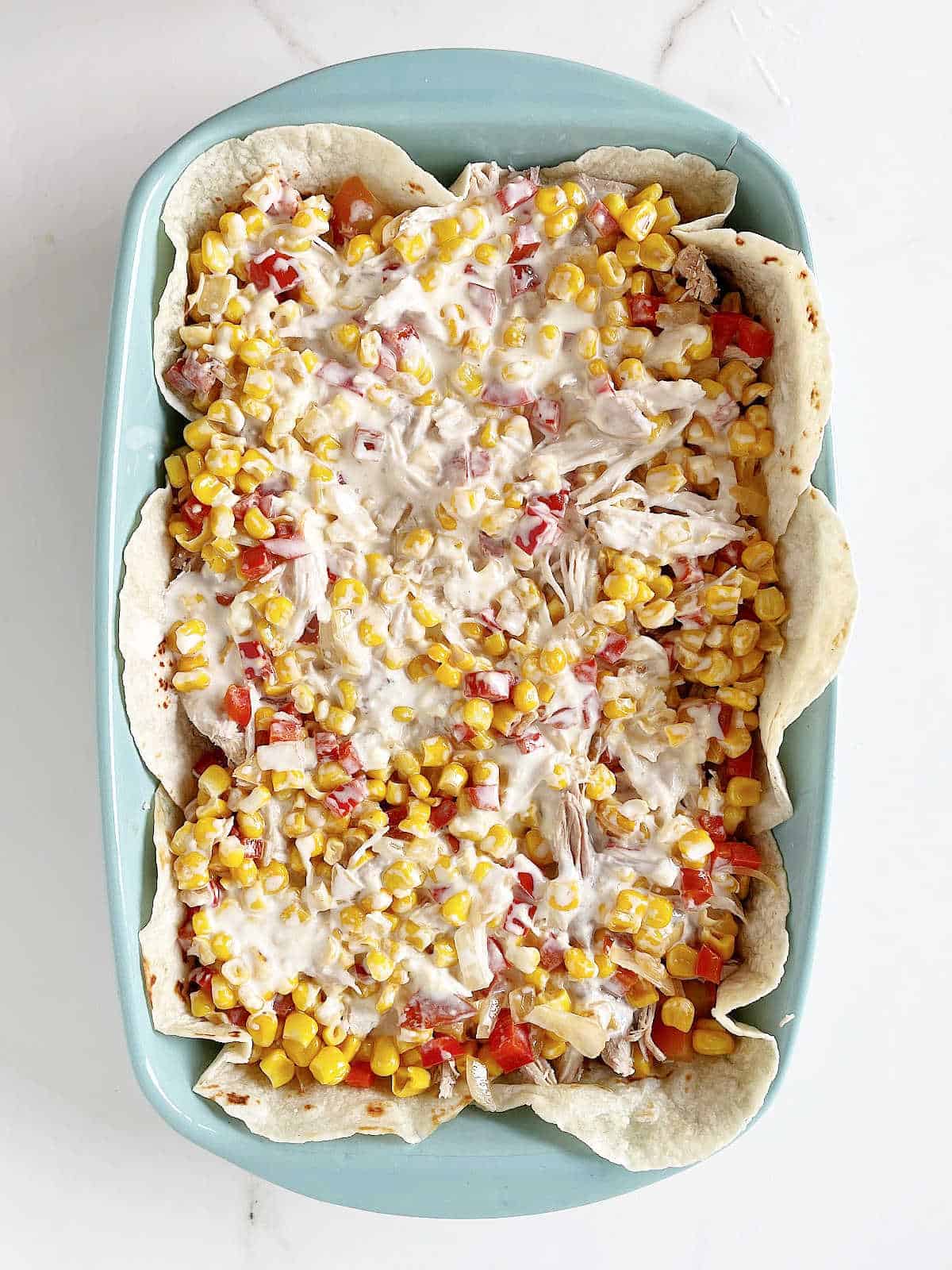 Corn, tomatoes, cream, and tortillas layered in a blue rectangular baking dish on a white marble surface.