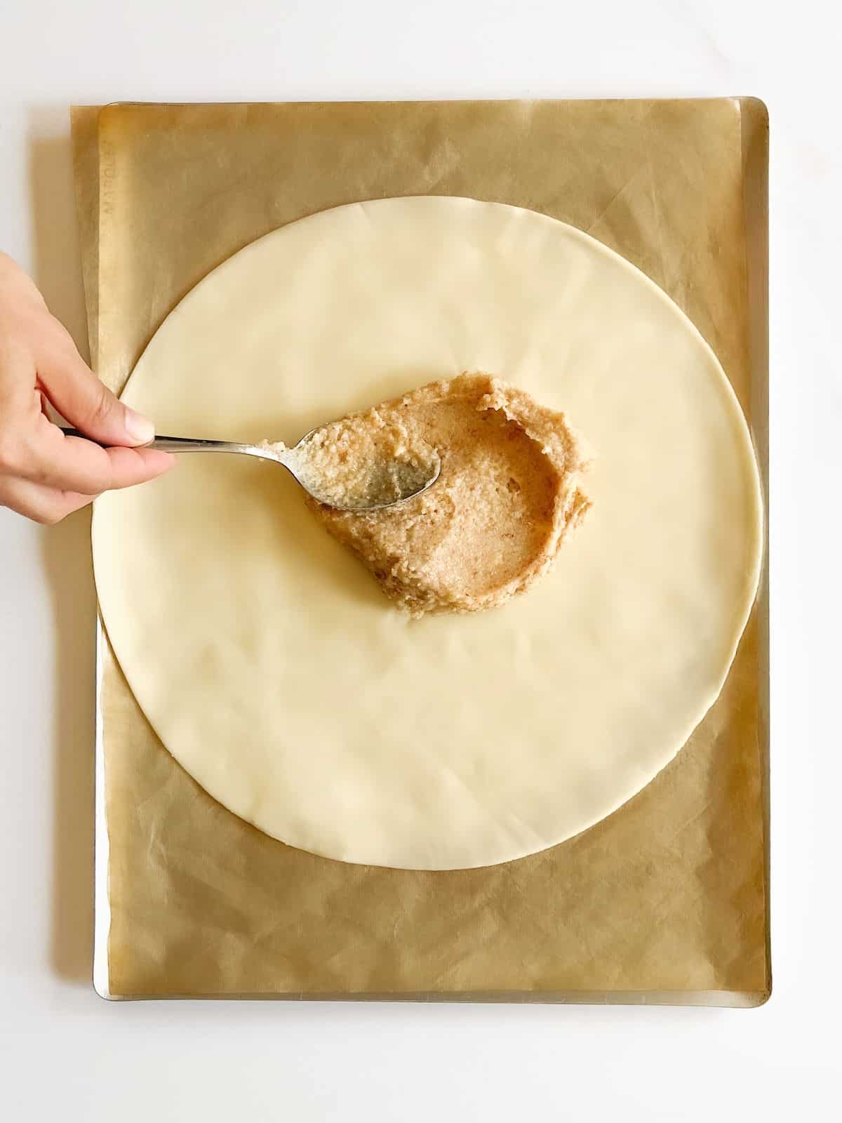 Spreading almond cream on a dough disc on beige parchment paper. White surface.