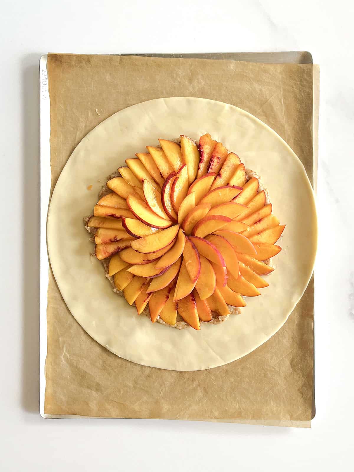 Sliced peaches making a circle on a round of pie crust. Beige parchment paper, white surface.